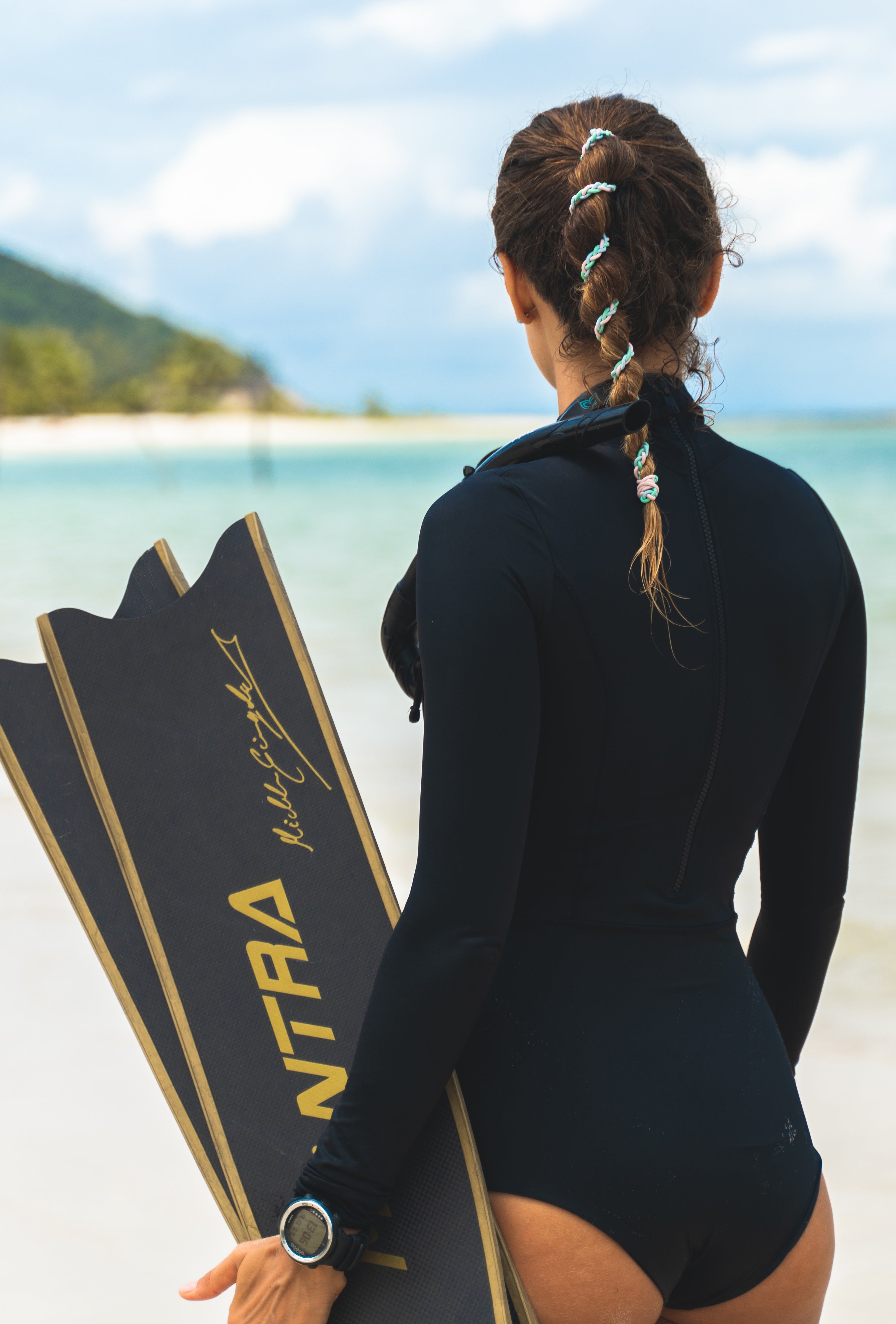 Woman in a wetsuit, holding diving fins, looking out at the crystal clear water of the Philippines with a Rip Tie hair tie holding her ponytail in place.